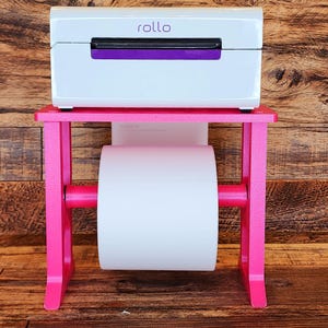 May include: A white label printer with a purple accent sits on a pink stand with a roll of white label paper. The printer has the word "rollo" printed on the front.