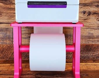 May include: A white label printer with a purple accent sits on a pink stand with a roll of white label paper. The printer has the word "rollo" printed on the front.