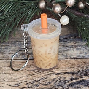 Puede incluir: Un llavero en miniatura que se asemeja a una bebida de t&eacute; de burbujas. El vaso de pl&aacute;stico transparente est&aacute; lleno de un l&iacute;quido marr&oacute;n claro y perlas de tapioca marrones. Tiene una tapa transparente con una pajita naranja y un llavero plateado.
