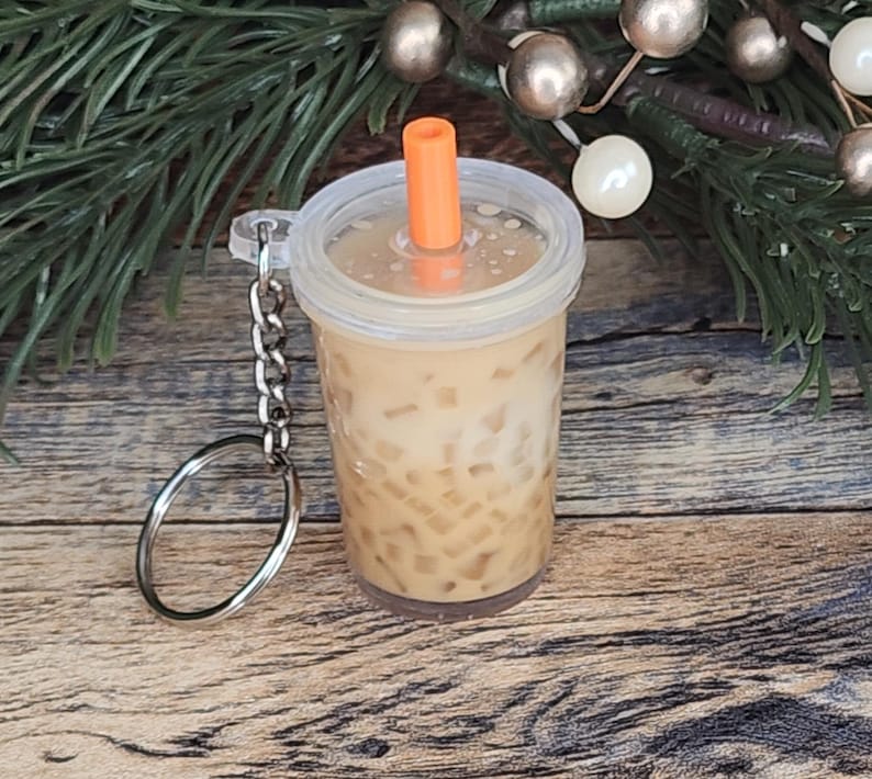 Puede incluir: Un llavero en miniatura que se asemeja a una bebida de t&eacute; de burbujas. El vaso de pl&aacute;stico transparente est&aacute; lleno de un l&iacute;quido marr&oacute;n claro y perlas de tapioca marrones. Tiene una tapa transparente con una pajita naranja y un llavero plateado.