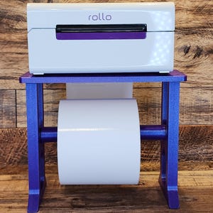 May include: A white label printer with the word "rollo" on the front, sitting on a purple metal stand with a roll of white label paper.