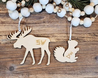 May include: Two wooden Christmas ornaments: a moose and a squirrel. The moose has the initials "SW" etched on its side, and the squirrel has "DW". Both are hanging from white ribbons, with a garland of white balls and gold beads in the background.