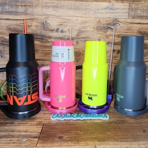 May include: Three stainless steel travel mugs with handles and lids. The mugs are in black, pink, and green. Each mug has a logo that says "Stanley" and is sitting on a matching colored coaster.