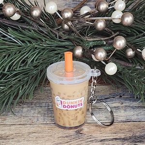 Puede incluir: Un llavero en miniatura de caf&eacute; helado de Dunkin' Donuts. El vaso de pl&aacute;stico transparente est&aacute; lleno de un l&iacute;quido marr&oacute;n claro y cubitos de hielo, coronado con una tapa transparente y una pajita naranja. El logotipo de Dunkin' Donuts es visible en el vaso.