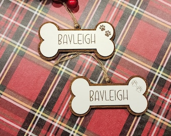 May include: Two white wooden bone-shaped ornaments with the name "BAYLEIGH" written on them. Each ornament has a small paw print design on the side.