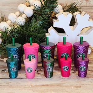 May include: A collection of eight miniature Starbucks tumblers with lids and straws. The tumblers are in various shades of green, pink, and purple with a Starbucks logo on each. The tumblers are arranged on a wooden surface with a white snowflake ornament and a white twig with white berries in the background.
