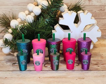 Op de afbeelding: Een collectie van vijf getextureerde Starbucks-bekers in teal, roze, donkergroen, felroze en paars, elk met een bijpassend rietje en logo. Daaronder bevinden zich vijf miniatuurversies van deze bekers bevestigd aan zilveren sleutelhangers.