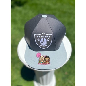 May include: A black and white paper hat with the word "RAIDERS" and a football player's face on the front. The hat has a gray brim and a white logo on the top. The hat is sitting on a white pedestal.