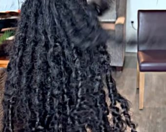 May include: A close-up of a long, thick, black, curly wig. The wig is styled in loose, textured curls and is hanging down in front of a light-colored background.