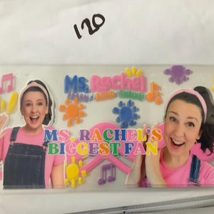 K&ouml;nnte beinhalten: Ein klarer Aufkleber mit Ms. Rachel und dem Text "Ms. Rachel's Biggest Fan". Das Design umfasst bunte Noten, Sonnen und Blumen. Ms. Rachel tr&auml;gt ein rosa Hemd und eine Jeans-Latzhose.