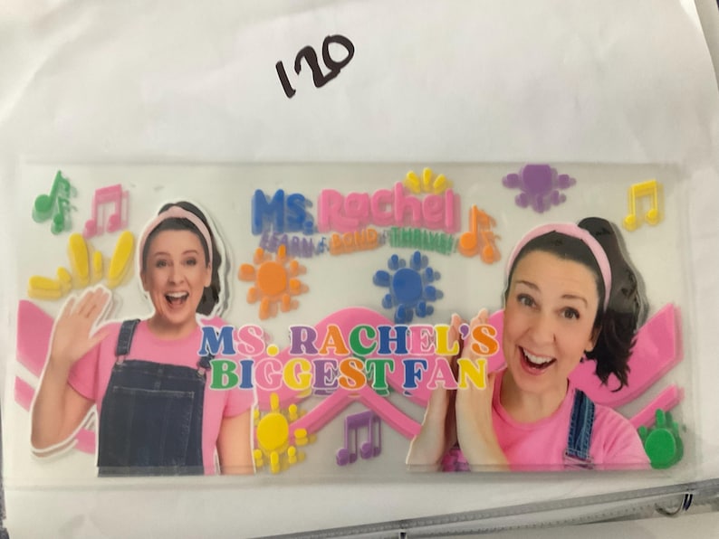 K&ouml;nnte beinhalten: Ein klarer Aufkleber mit Ms. Rachel und dem Text "Ms. Rachel's Biggest Fan". Das Design umfasst bunte Noten, Sonnen und Blumen. Ms. Rachel tr&auml;gt ein rosa Hemd und eine Jeans-Latzhose.