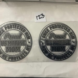 K&ouml;nnte beinhalten: Zwei runde Aufkleber mit dem Text "I ONCE PROTECTED HIM, NOW HE PROTECTS ME", der "Proud US MARINE MOM" umkreist. Das Design ist in Grau- und Wei&szlig;t&ouml;nen gehalten. Ein kleines wei&szlig;es Etikett mit der Nummer "123" ist sichtbar.