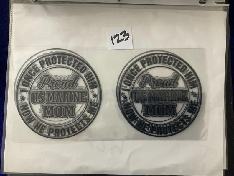 K&ouml;nnte beinhalten: Zwei runde Aufkleber mit dem Text "I ONCE PROTECTED HIM, NOW HE PROTECTS ME", der "Proud US MARINE MOM" umkreist. Das Design ist in Grau- und Wei&szlig;t&ouml;nen gehalten. Ein kleines wei&szlig;es Etikett mit der Nummer "123" ist sichtbar.
