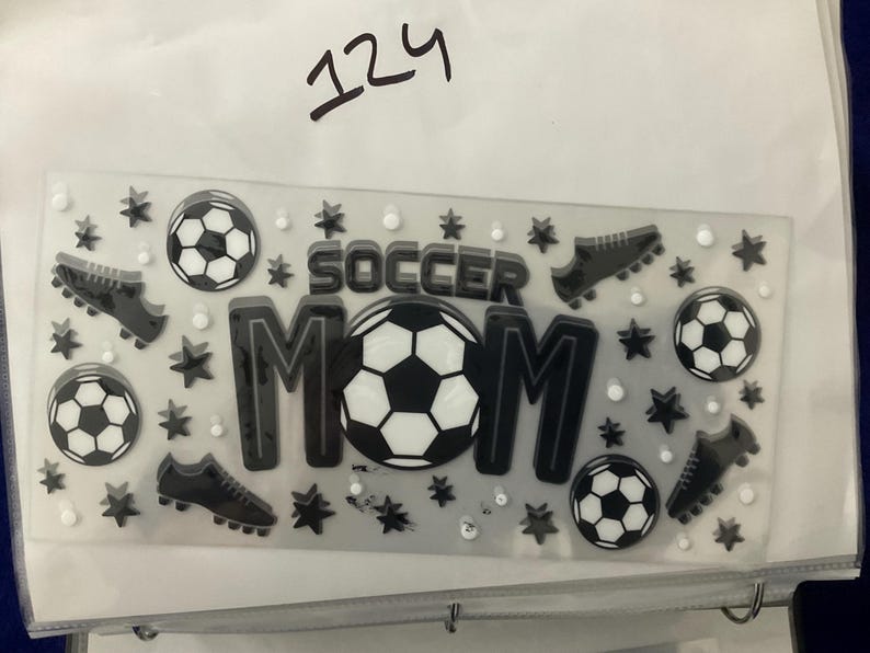 K&ouml;nnte beinhalten: Ein klarer Aufkleberbogen mit Fu&szlig;ball-Motiv. Das Design zeigt die Worte "SOCCER MOM" in Schwarz und Wei&szlig;, Fu&szlig;b&auml;lle, Fu&szlig;ballschuhe, Sterne und kleine wei&szlig;e Kreise. Die Nummer "124" ist handschriftlich oben.