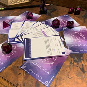 May include: A set of purple and white warlock spell cards with a purple and gold dice on top of one of the cards. The cards have a geometric design and the text "WARLOCK Spell Card" on them.