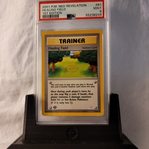 May include: A Pokemon Trading Card Game card featuring Healing Field, a Stadium card from the Neo Revelation set. The card is in a protective case with a PSA grading label. The card features a green and brown illustration of a forest clearing with a Pokemon symbol in the center.