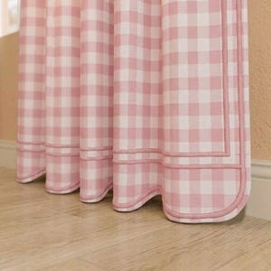 Può includere: Tende a quadretti rosa e bianche con bordo e finiture decorative. Le tende sono appese davanti a un muro colorato chiaro e a un pavimento in legno. Il tessuto ha un'estetica classica e country chic.