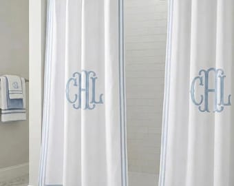 Może przedstawiać: Biała zasłona prysznicowa z wyhaftowanym jasnoniebieskim monogramem z literami CHL, ozdobiona podwójnymi jasnoniebieskimi paskami na brzegach i wykończona falowanym dołem, zawieszona na metalowym drążku z kółkami w łazience.