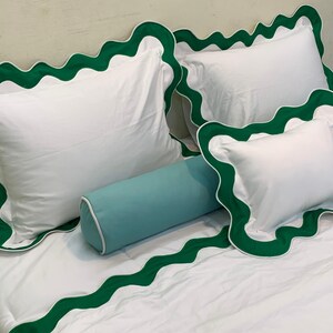 May include: White bedding set with green scalloped trim. The set includes two square pillows, a rectangular pillow, and a bolster pillow. The green trim is a vibrant accent against the white fabric. The bedding is neatly arranged on a bed.