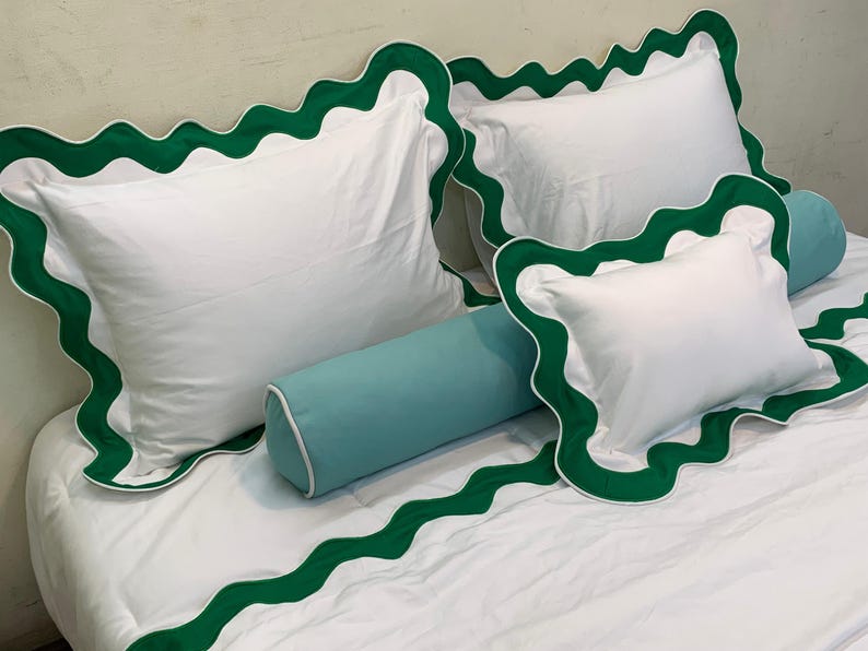 May include: White bedding set with green scalloped trim. The set includes two square pillows, a rectangular pillow, and a bolster pillow. The green trim is a vibrant accent against the white fabric. The bedding is neatly arranged on a bed.