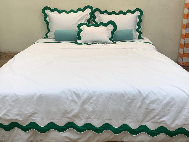 May include: White bed linens with green scalloped trim and matching pillowcases. The bed is made with a white comforter, two standard pillowcases, a decorative pillow, and a bolster pillow. The bed set is a solid white color.