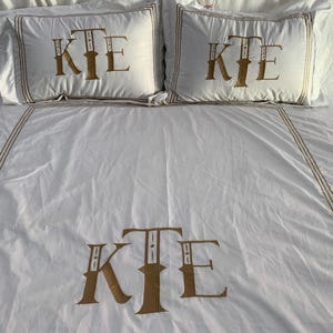 May include: White bedding set featuring embroidered gold initials "KTE" on the pillowcases and duvet cover. The set includes two pillowcases and a duvet cover, with gold trim.