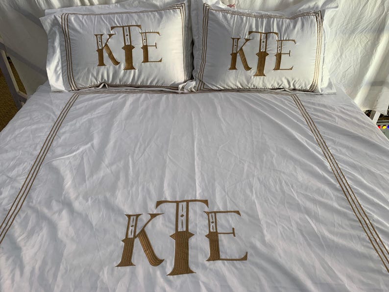 May include: White bedding set featuring embroidered gold initials "KTE" on the pillowcases and duvet cover. The set includes two pillowcases and a duvet cover, with gold trim.