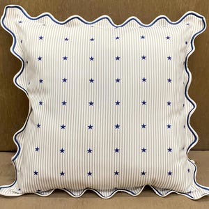 Op de afbeelding: Een vierkant decoratief kussen met een witte achtergrond met verticale blauwe strepen en blauwe sterren. Het kussen heeft een geschulpte rand met een blauwe bies.