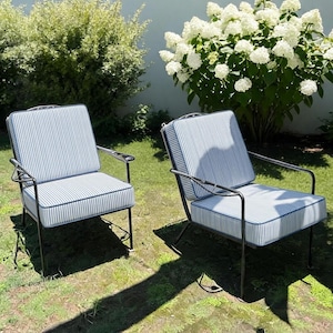 K&ouml;nnte beinhalten: Zwei Outdoor-Sessel mit schwarzen Metallrahmen und blau-wei&szlig; gestreiften Kissen. Die St&uuml;hle stehen auf einem Rasen, im Hintergrund ein wei&szlig; bl&uuml;hender Busch.