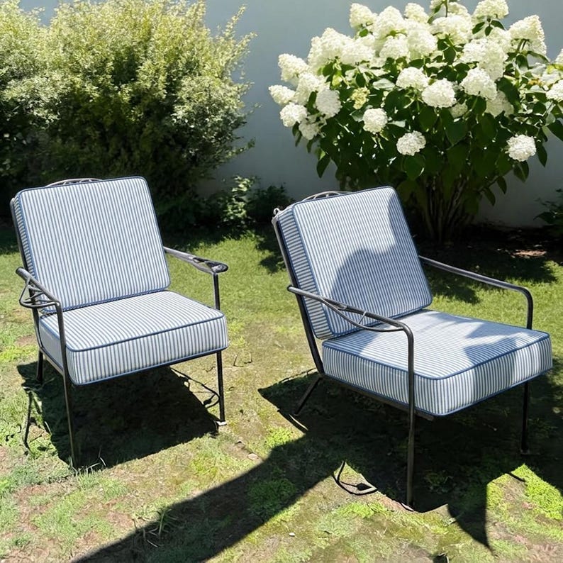 K&ouml;nnte beinhalten: Zwei Outdoor-Sessel mit schwarzen Metallrahmen und blau-wei&szlig; gestreiften Kissen. Die St&uuml;hle stehen auf einem Rasen, im Hintergrund ein wei&szlig; bl&uuml;hender Busch.