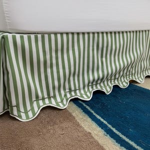 Pode incluir: Uma saia de cama com listras verticais verdes e brancas. A saia tem uma borda ondulada com acabamento branco. A saia de cama é feita de tecido e foi projetada para cobrir a base da cama.