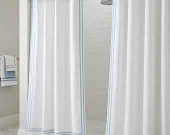 May include: White shower curtains with a light blue border and trim hang from a silver rod. The curtains are in front of a white tiled shower. Matching towels are on a nearby rack.