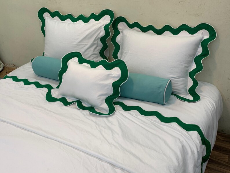May include: White bedding set with green scalloped trim. The set includes two square pillows, a smaller square pillow, and a bolster pillow in a light blue color. The bedspread also features the green scalloped trim.