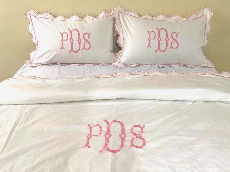 May include: White bedding set with pink scalloped edges and pink embroidered initials "PDS" on the pillowcases and duvet cover.