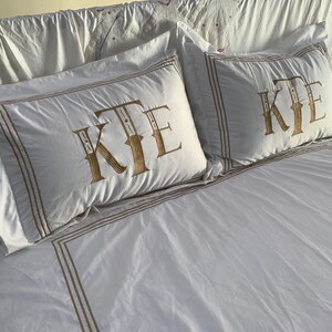 May include: White bedding set featuring two pillowcases with a gold-coloured border and monogram. The monogram on the pillowcases reads "KTE". The bedsheet also has a gold-coloured border.