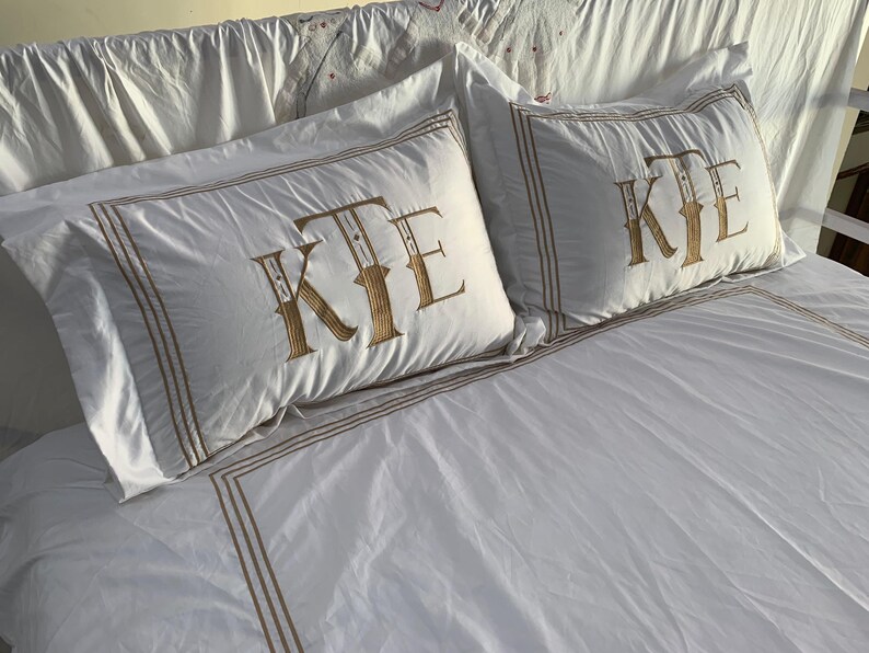 May include: White bedding set featuring two pillowcases with a gold-coloured border and monogram. The monogram on the pillowcases reads "KTE". The bedsheet also has a gold-coloured border.