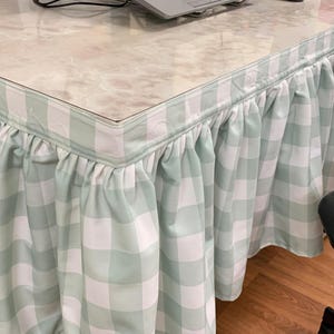 以下が含まれることがあります： 薄緑と白のチェック柄のデスクスカート。スカートは布製で、フリルの縁が付いています。デスクはマーブル模様の天板です。