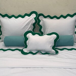 May include: White bedding set with green scalloped trim. The set includes two square pillows, a rectangular bolster pillow, and a smaller square pillow. The bedspread also features the green scalloped trim. The bedding is neatly arranged on a bed.