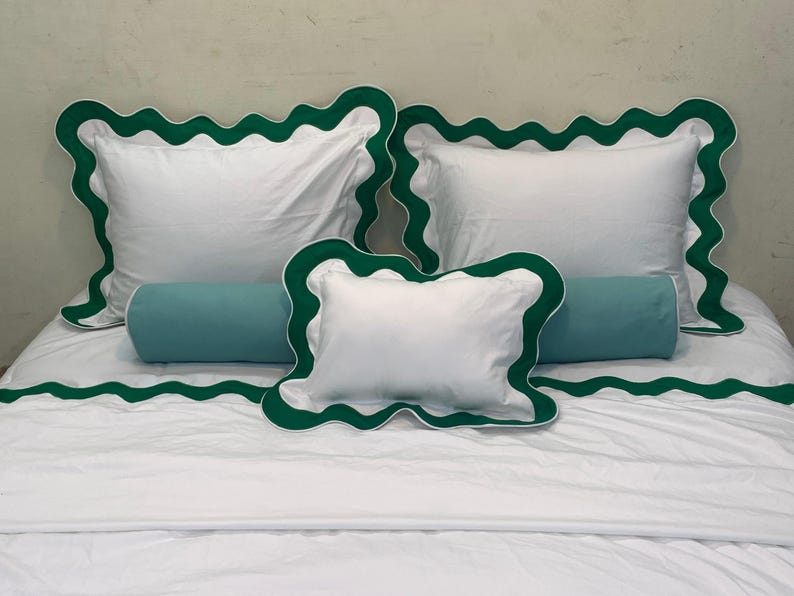 May include: White bedding set with green scalloped trim. The set includes two square pillows, a rectangular bolster pillow, and a smaller square pillow. The bedspread also features the green scalloped trim. The bedding is neatly arranged on a bed.