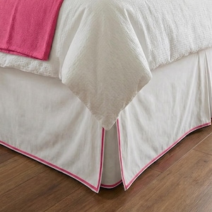 May include: White bed skirt with a pink trim. The bed skirt is made of a crisp, white fabric and features a tailored design. A pink blanket is visible on the bed.
