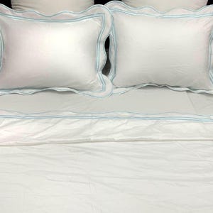 Puede incluir: Sábanas de cama blancas con ribete azul claro. La cama está hecha con una funda nórdica blanca y dos fundas de almohada blancas con bordes festoneados y ribete azul claro.