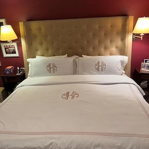 May include: A bed with a beige upholstered headboard and white bedding. The bedding includes a duvet cover and two pillowcases, all embroidered with a monogram. The duvet cover has a thin red stripe near the bottom.