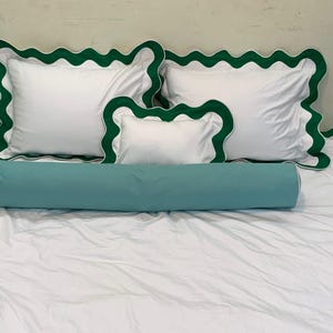 May include: White bedding set with green scalloped trim. Includes two square pillows, a smaller rectangular pillow, and a cylindrical bolster pillow in a teal color. The bed is covered with a white sheet.