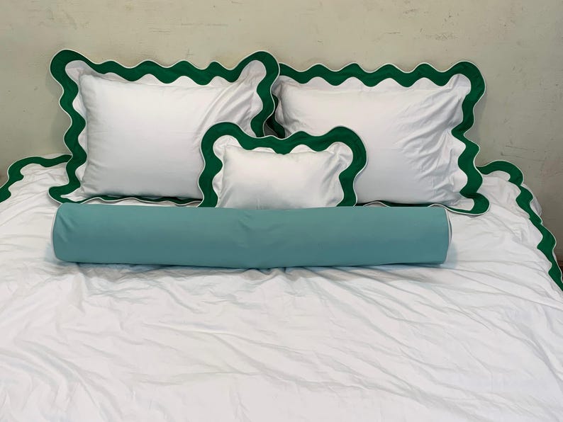 May include: White bedding set with green scalloped trim. Includes two square pillows, a smaller rectangular pillow, and a cylindrical bolster pillow in a teal color. The bed is covered with a white sheet.