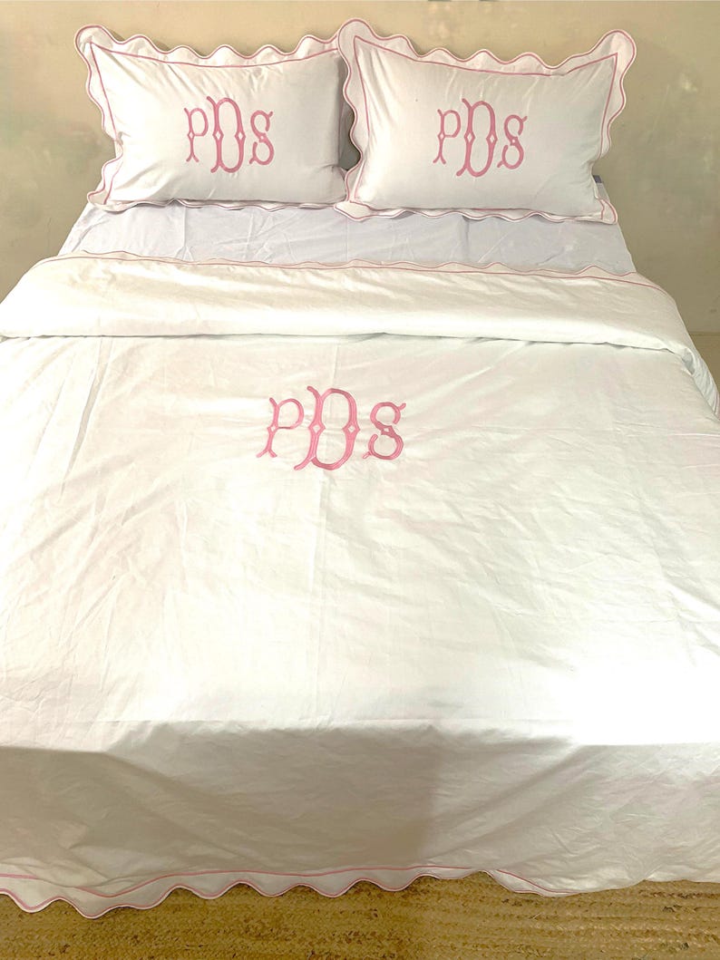 May include: White bedding set with pink scalloped edges and pink embroidered initials "PDS" on the pillowcases and duvet cover.