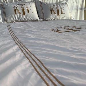 May include: White bed linen set featuring gold embroidered initials on the pillowcases and duvet cover. The pillowcases have a gold border. The duvet cover has gold stripes. The bed is made up.