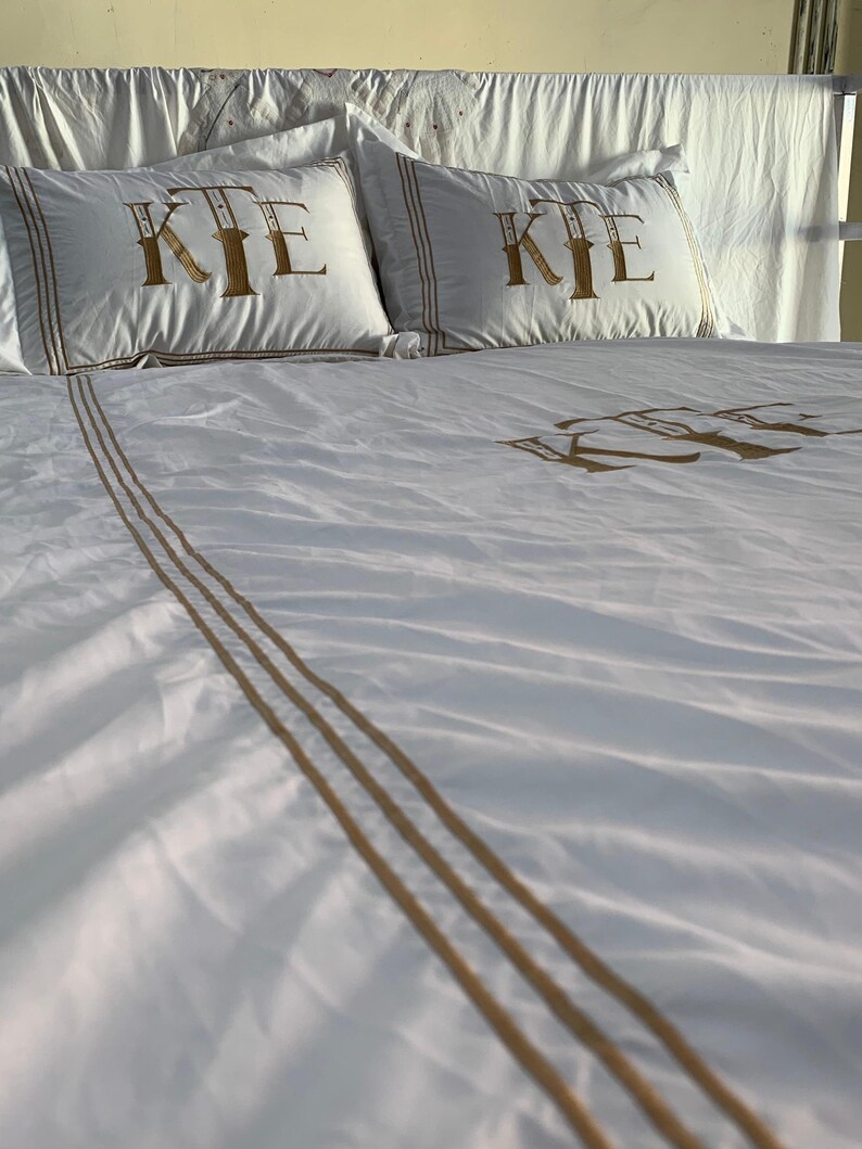 May include: White bed linen set featuring gold embroidered initials on the pillowcases and duvet cover. The pillowcases have a gold border. The duvet cover has gold stripes. The bed is made up.