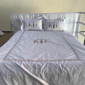 May include: White bed linen set featuring gold embroidered trim and the monogram "KIE" on the pillowcases and duvet cover. The set includes two pillowcases and a duvet cover, offering a coordinated aesthetic for the bed.