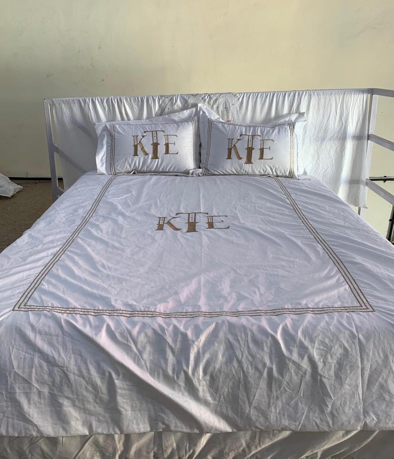 May include: White bed linen set featuring gold embroidered trim and the monogram "KIE" on the pillowcases and duvet cover. The set includes two pillowcases and a duvet cover, offering a coordinated aesthetic for the bed.