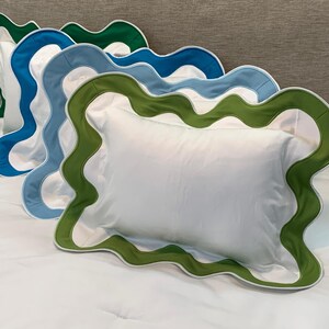 May include: Four white rectangular pillows with scalloped edges in green, blue, and white are arranged on a white bed. The pillows have a solid white center and a contrasting border. The pillows are placed on a white bedspread.